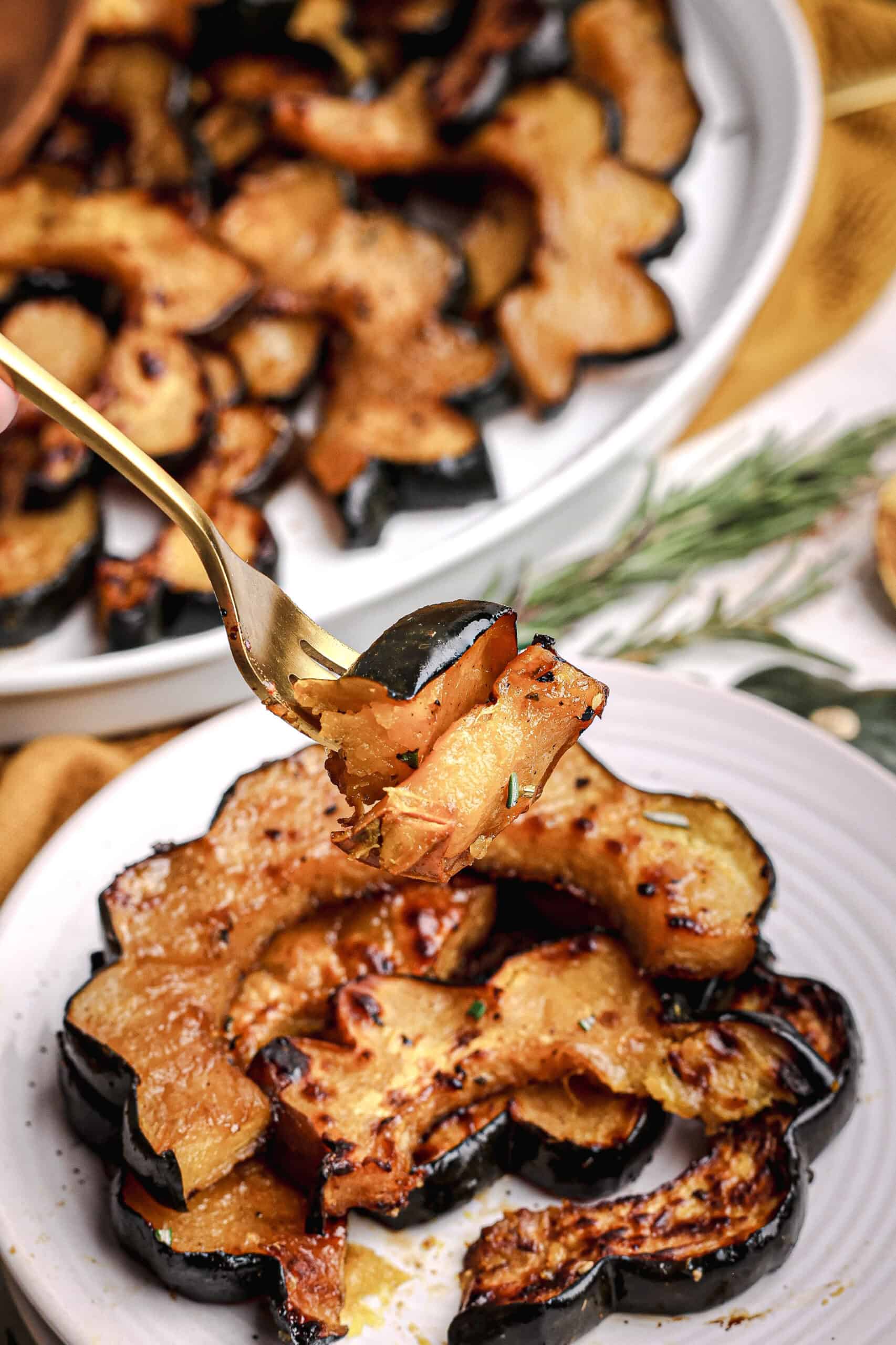 Close-up of a fork lifting a caramelized slice of maple roasted acorn squash from a plate, showing glossy edges and soft golden interior with rosemary in the background.