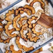 Maple roasted acorn squash slices spread across a parchment-lined baking sheet, surrounded by warm gold linens, rosemary, and kitchen utensils.