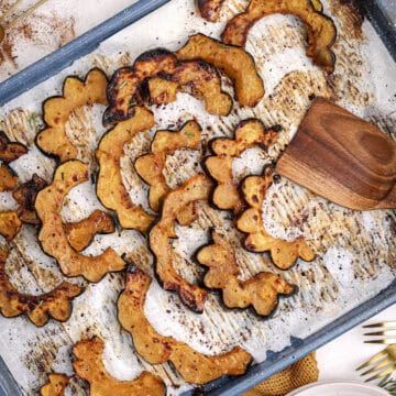 Maple roasted acorn squash slices spread across a parchment-lined baking sheet, surrounded by warm gold linens, rosemary, and kitchen utensils.
