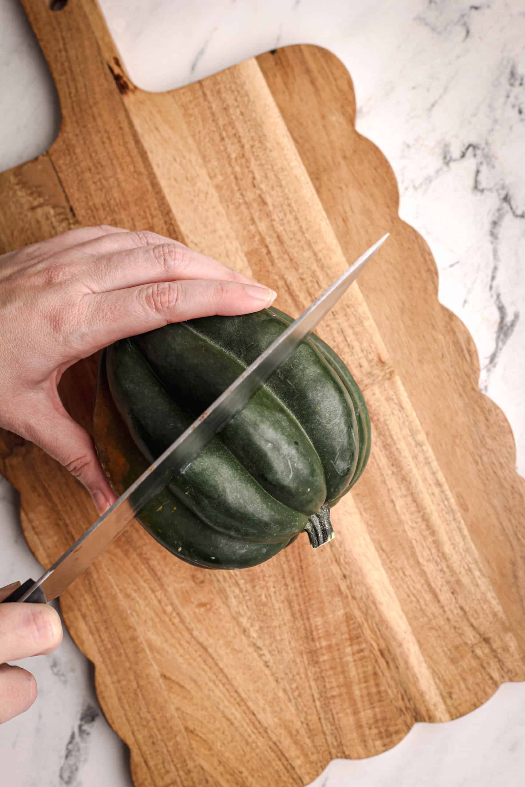 A hand holds an acorn squash steady on a wooden cutting board while a large knife begins slicing through the center.