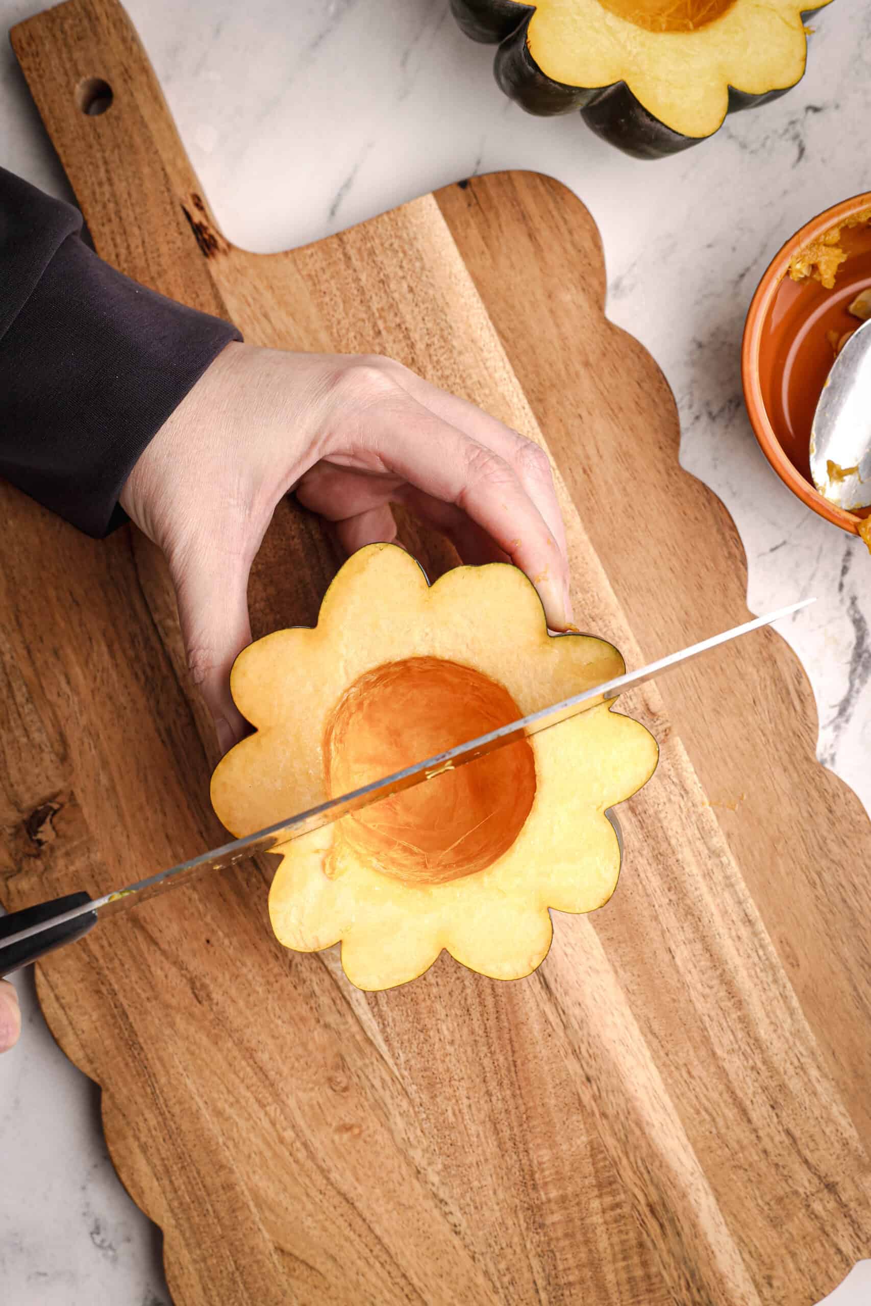 A hand stabilizing a cleaned acorn squash half while a knife slices it into sections on a wooden cutting board.