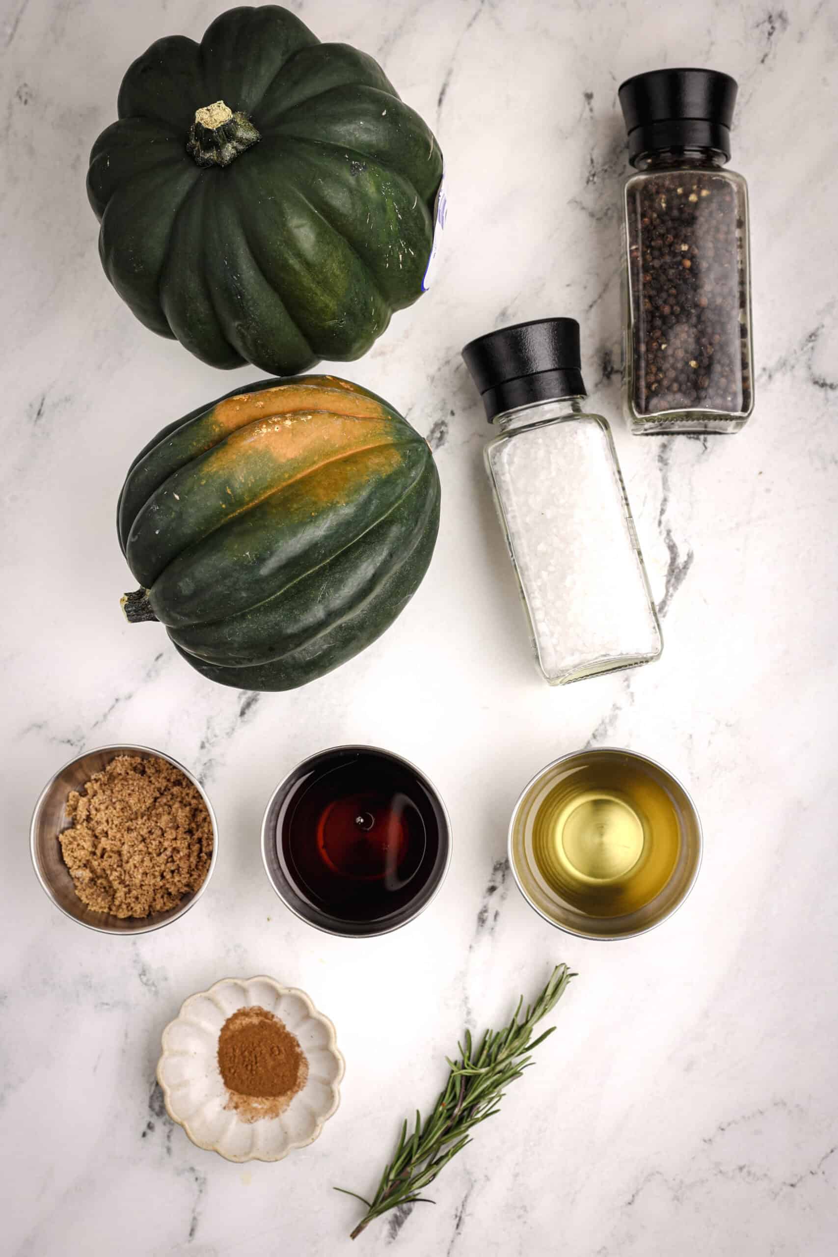 Overhead shot of whole acorn squash, bowls of brown sugar, maple syrup, and oil, along with salt, pepper, cinnamon, and a sprig of rosemary arranged on a white marble surface.