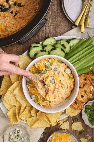 Bowl of buffalo chicken dip surrounded by tortilla chips, sliced cucumbers, celery sticks, and pretzel crisps, with a wooden spoon scooping into the dip.