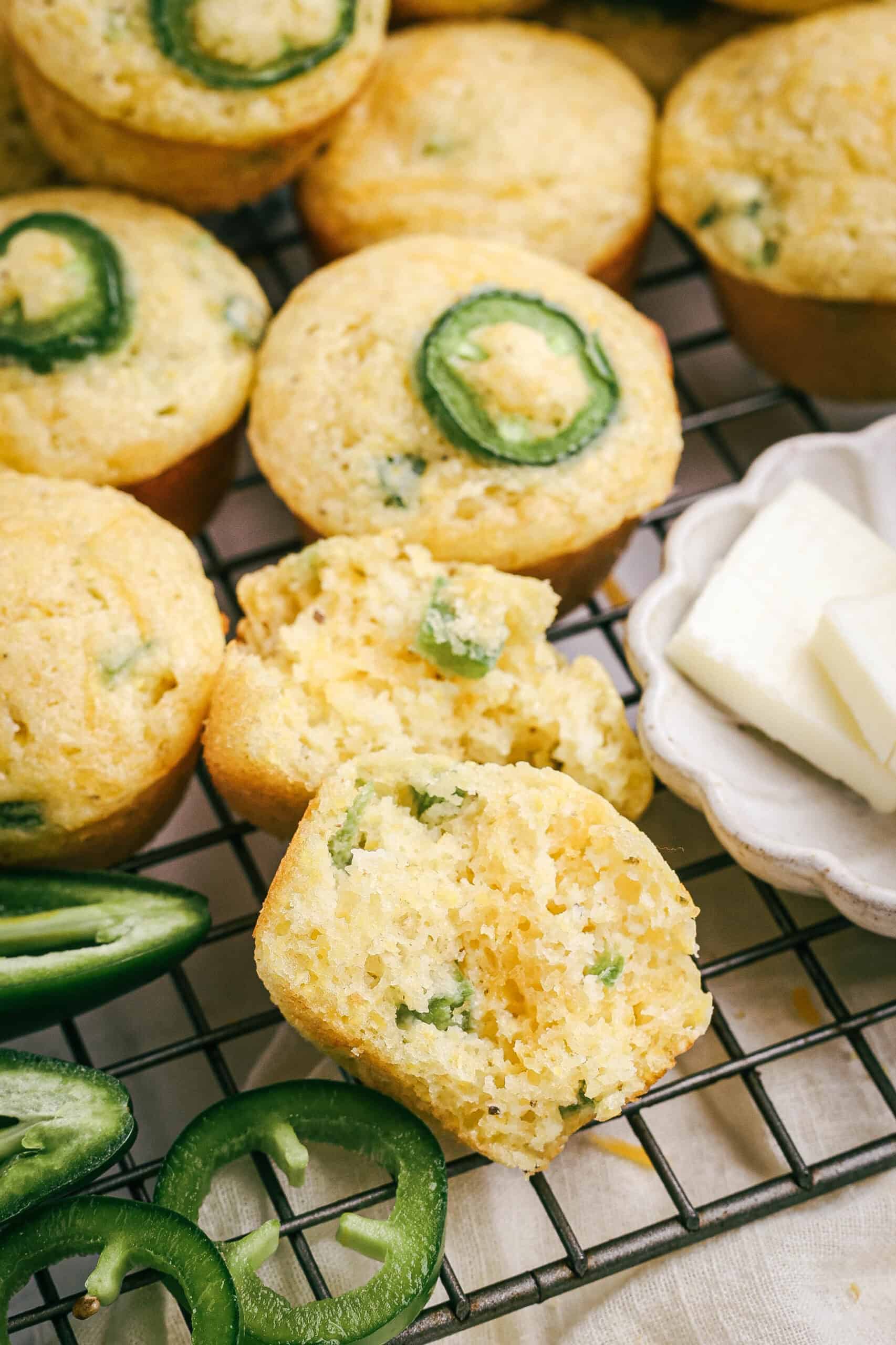 Golden mini jalapeño cheddar cornbread muffins cooling on a wire rack, one split open to reveal a moist, cheesy center. Fresh jalapeño slices and butter are placed beside for serving.