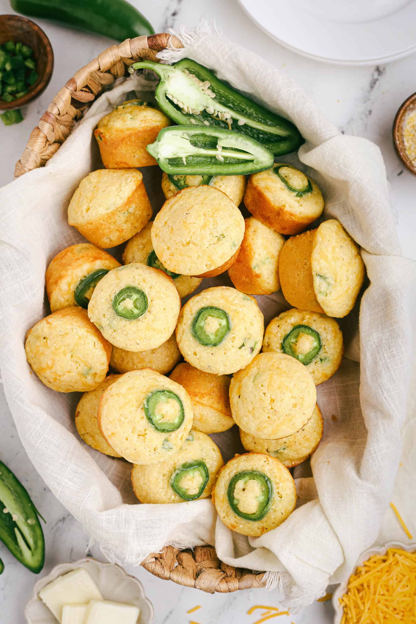 Basket lined with linen cloth filled with freshly baked mini jalapeño cheddar cornbread muffins, each topped with a jalapeño slice, surrounded by shredded cheese and fresh peppers.