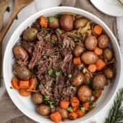 Shredded slow cooker pot roast served in a large white bowl with carrots, red potatoes, and onions, garnished with fresh herbs.