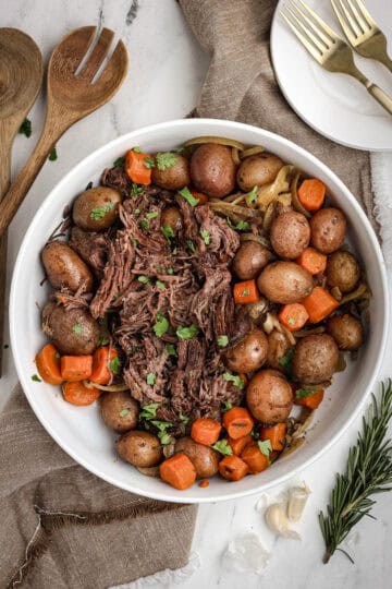 Shredded slow cooker pot roast served in a large white bowl with carrots, red potatoes, and onions, garnished with fresh herbs.