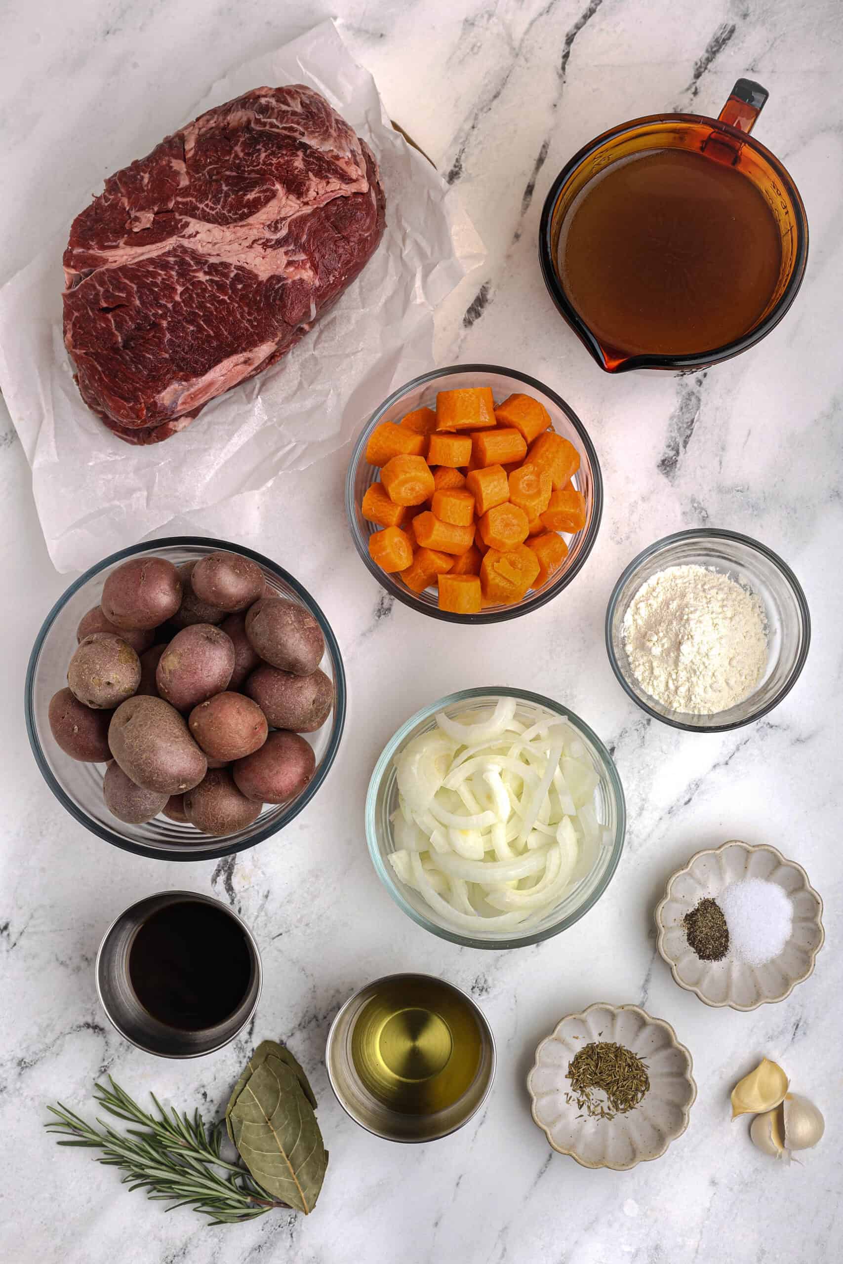 Ingredients for slow cooker pot roast laid out on a marble surface, including a chuck roast, carrots, potatoes, onions, beef broth, flour, garlic, olive oil, and herbs.