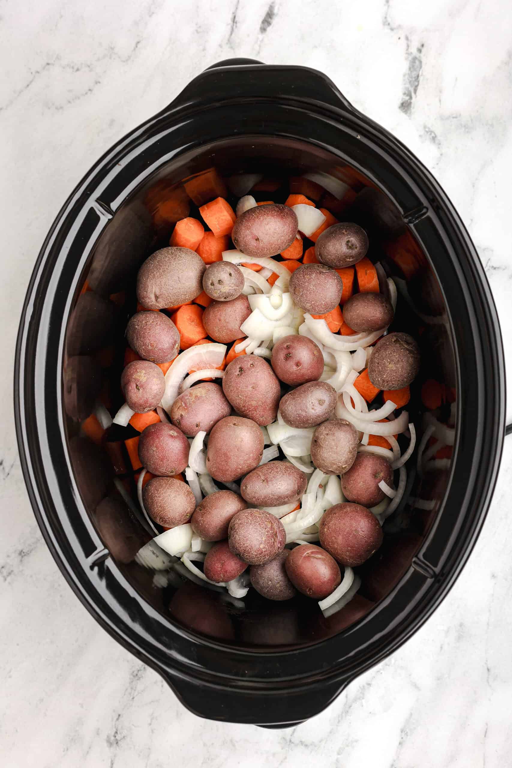 Sliced carrots, red potatoes, and onions layered in the bottom of a slow cooker, ready for pot roast.