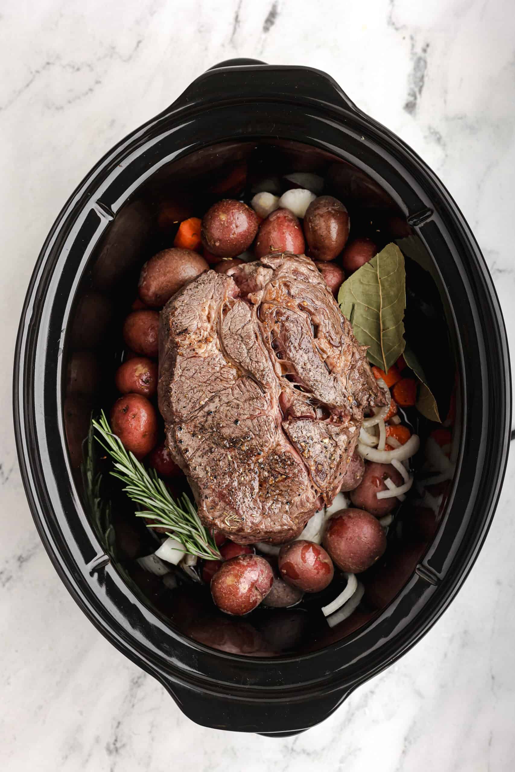 Sear-browned beef roast placed over vegetables in a slow cooker with rosemary and bay leaves before cooking.