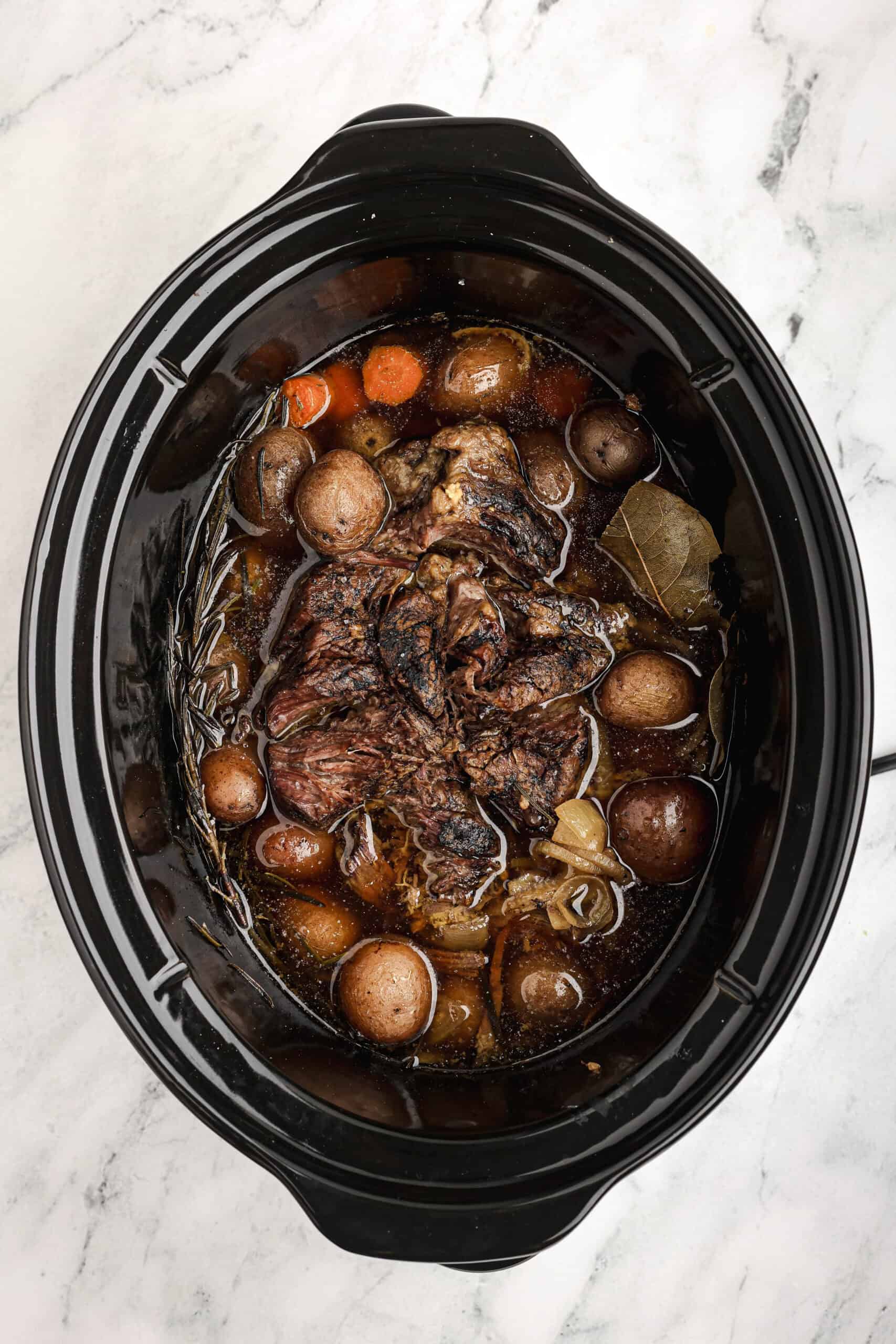 Tender beef chuck roast, carrots, and red potatoes simmering in rich broth inside a slow cooker after cooking.