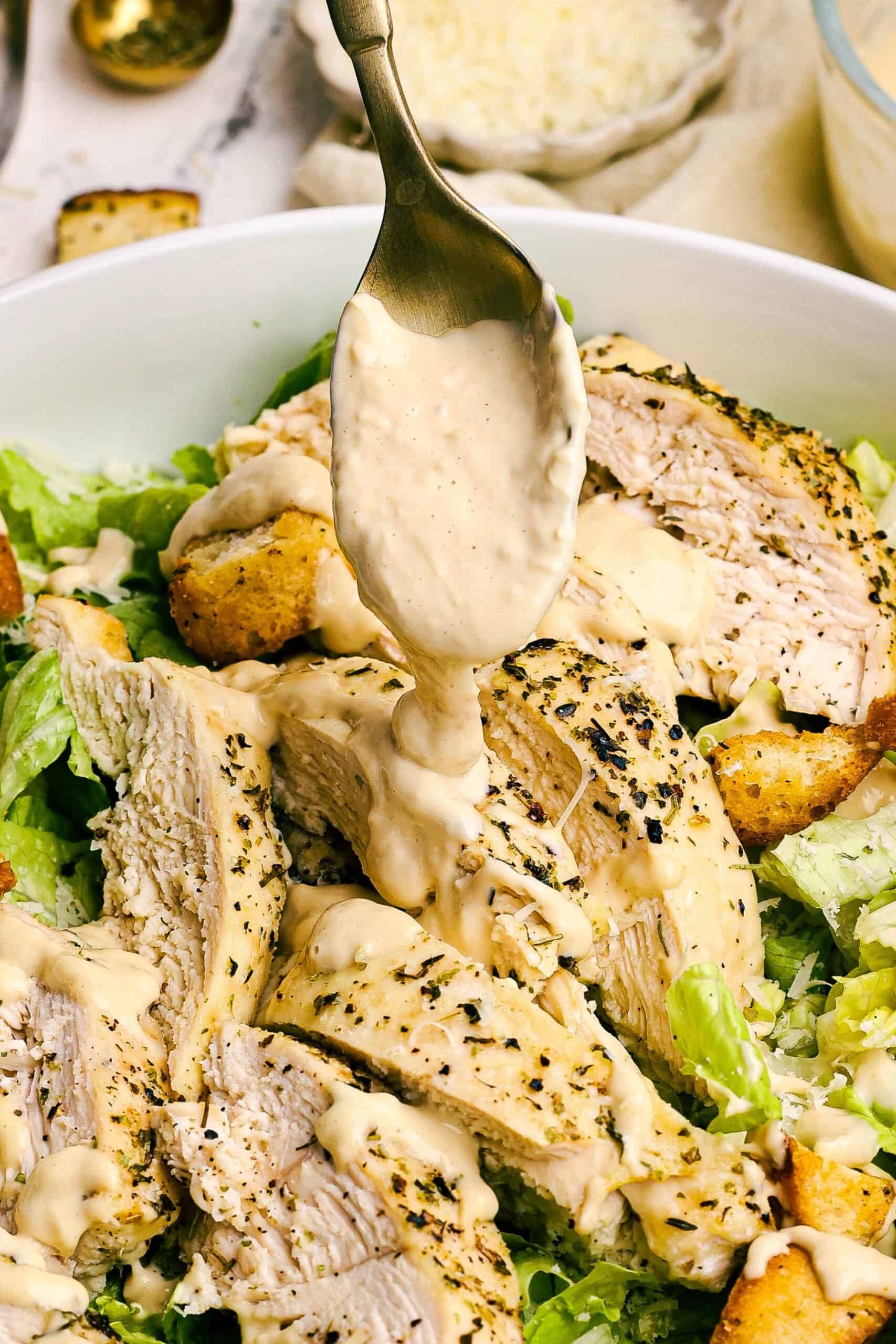 Close-up of creamy Caesar dressing being spooned over sliced chicken and romaine lettuce in a Caesar salad