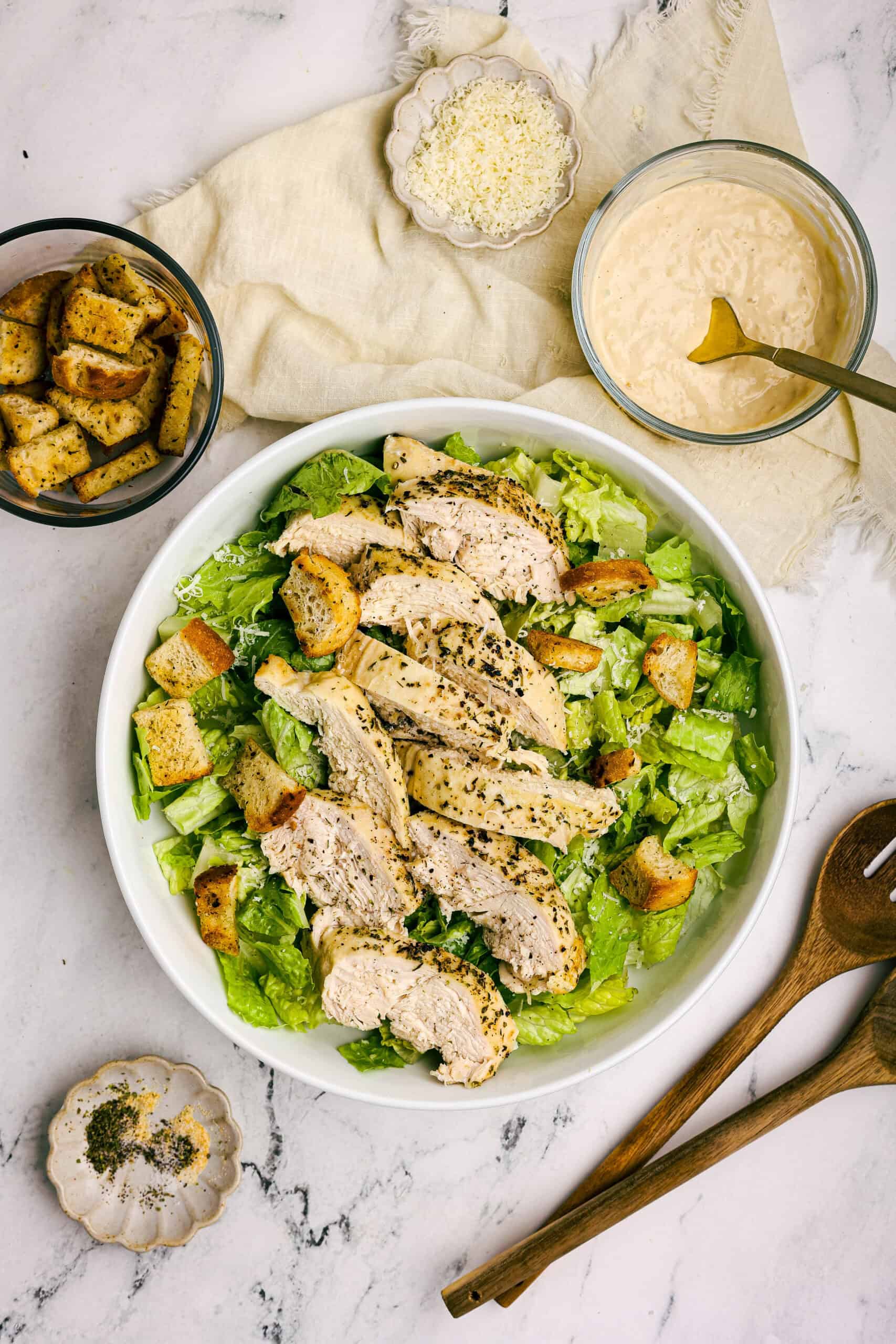 Chicken Caesar salad with homemade croutons, sliced chicken breast, and creamy dressing surrounded by salad utensils and ingredients
