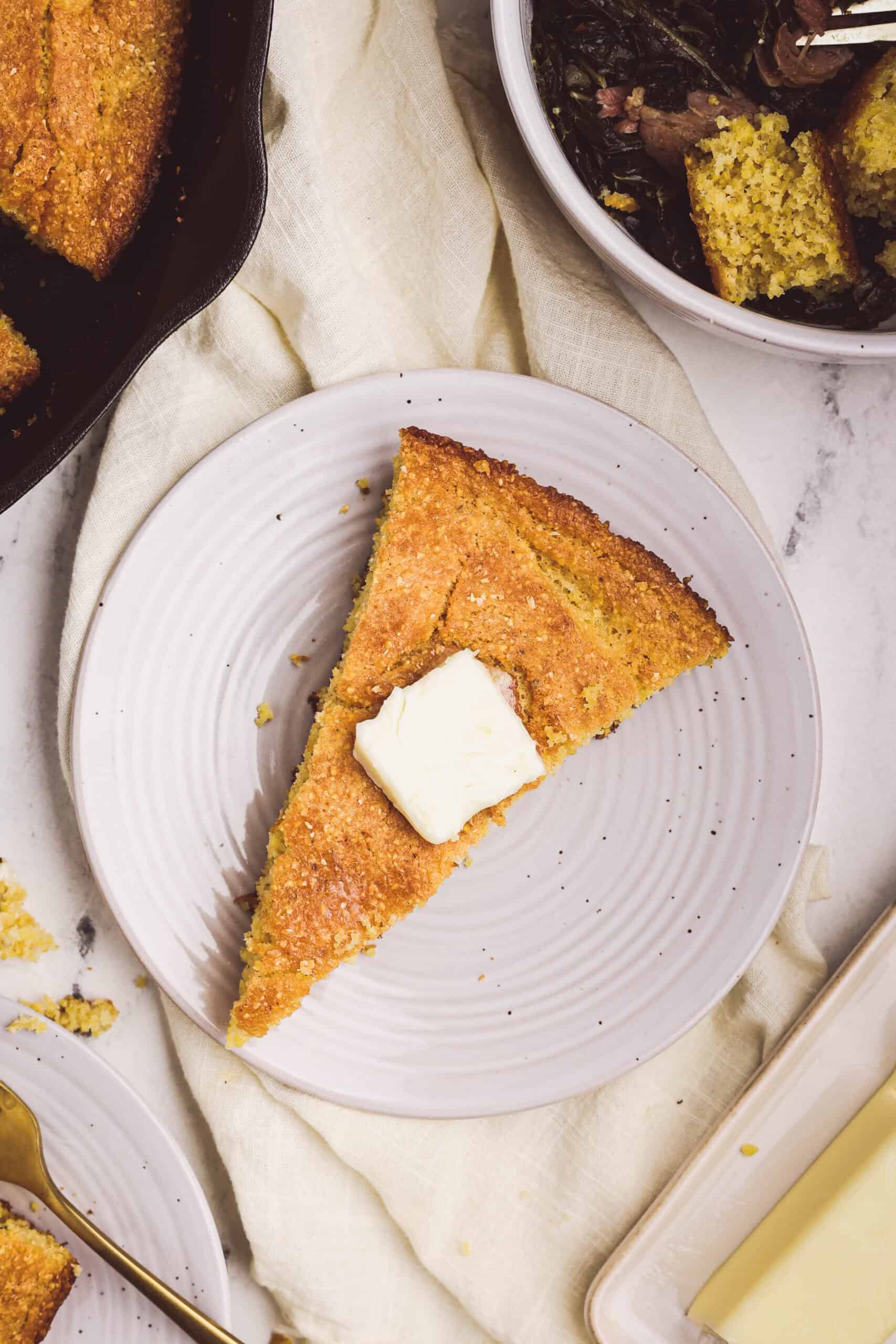 Slice of cast iron cornbread served on a plate with butter, alongside collard greens.