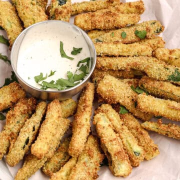 Plate of crispy baked zucchini fries served with a bowl of creamy dipping sauce, garnished with fresh herbs.