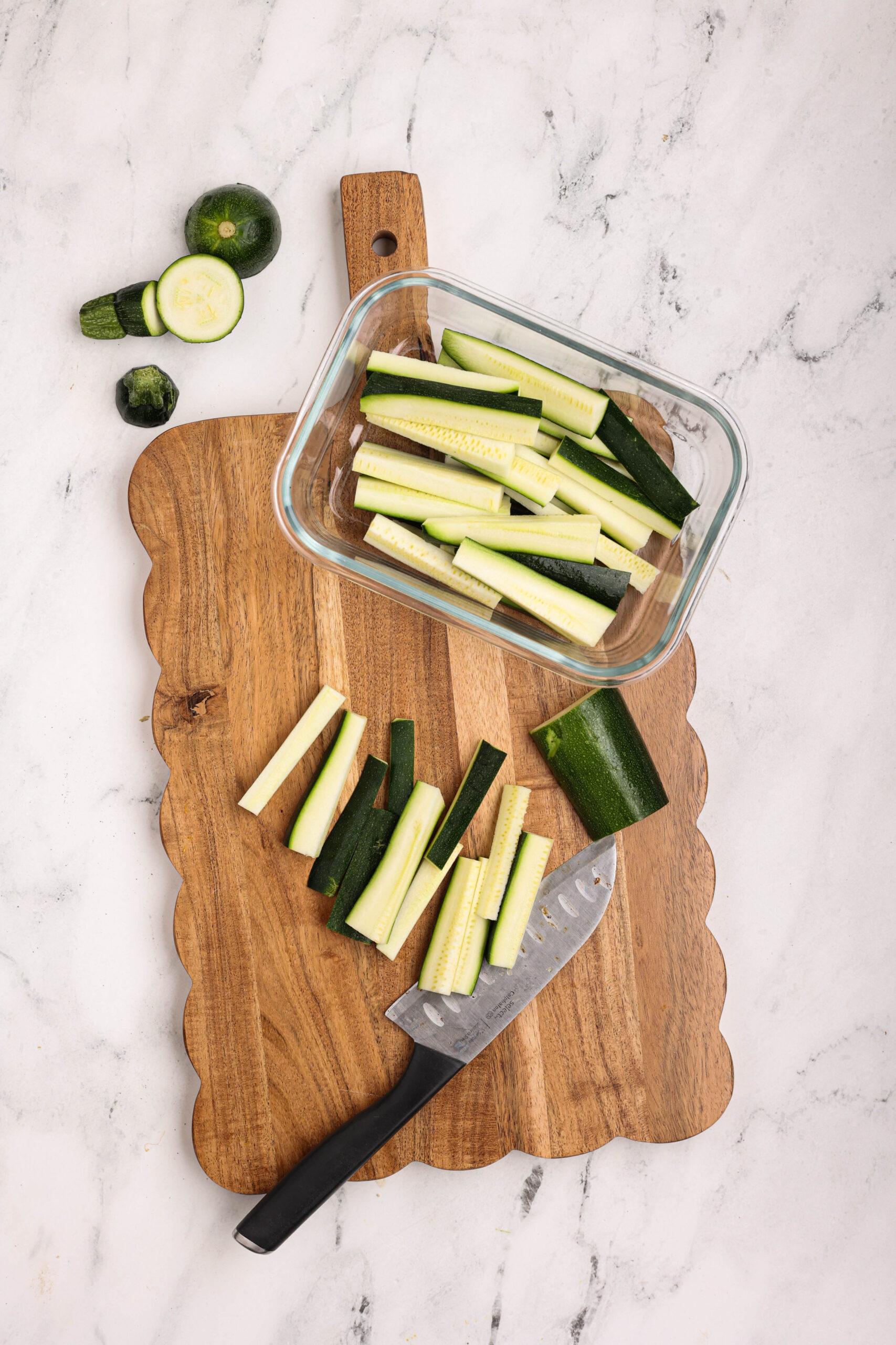Raw zucchini cut into fry-shaped sticks arranged on a wooden cutting board with a chef’s knife.