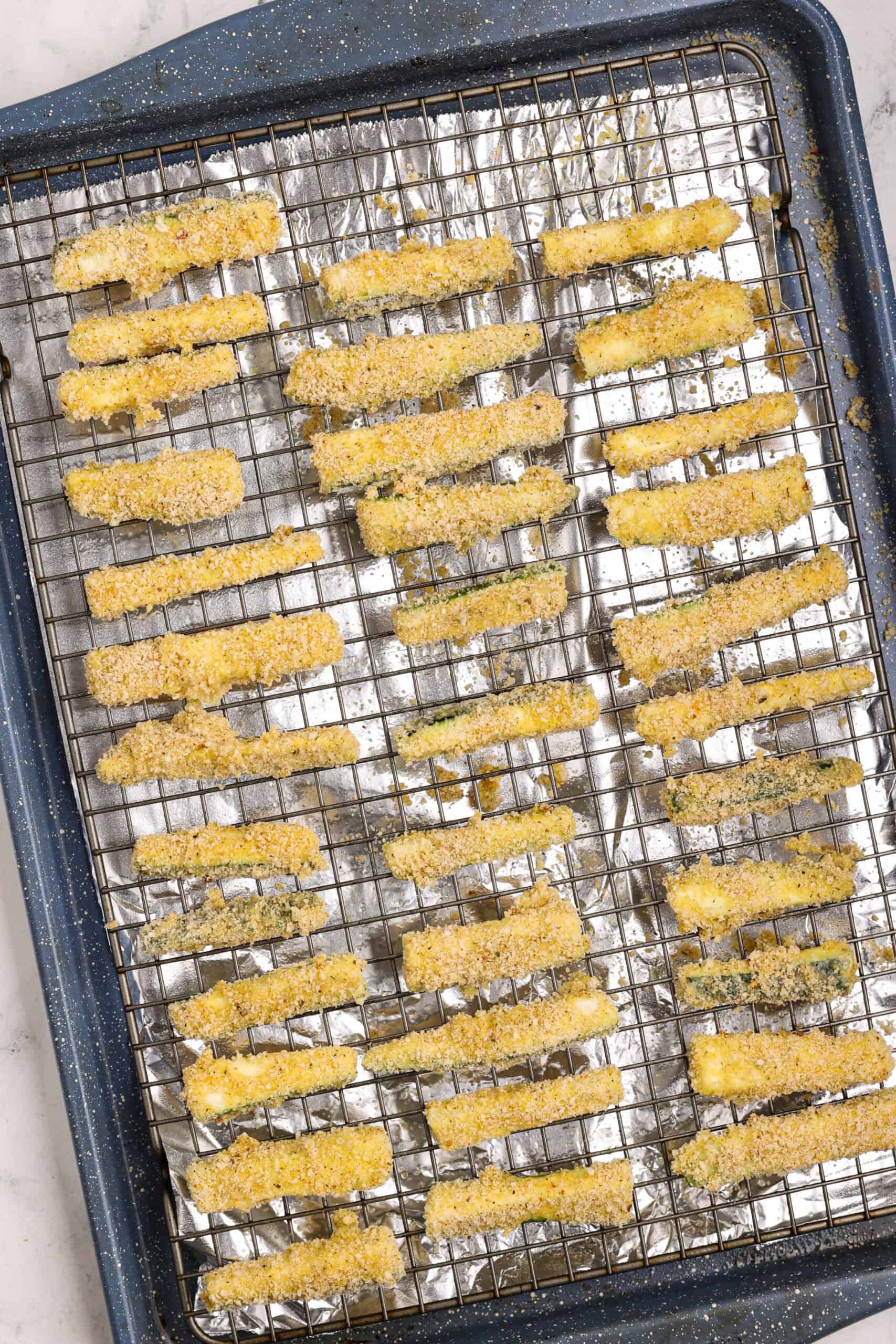 Breaded zucchini fries arranged on a greased wire rack, ready to be baked in the oven.