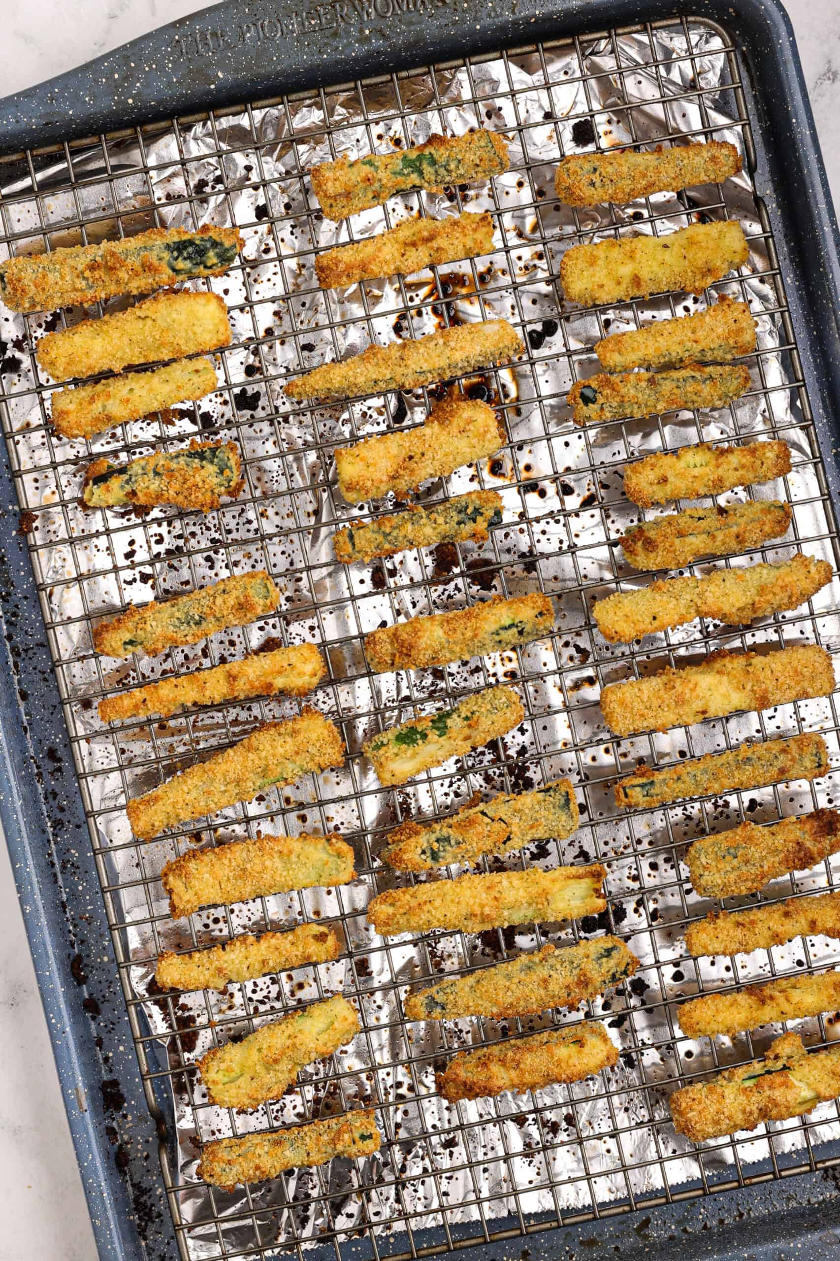 Crispy baked zucchini fries arranged on a wire rack over a foil-lined baking sheet, showing even browning and golden crumb coating.